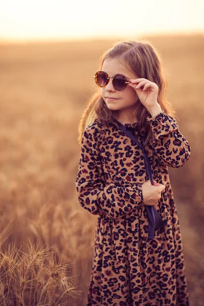 Модна фотографія маленької дівчинки в сукні для друку леопардів, сонцезахисних окулярах та солом'яному капелюсі на вечірньому пшеничному полі — стокове фото