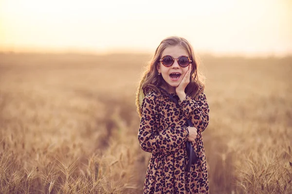 Mode foto av en liten flicka i leopard print klänning, solglasögon och halm hatt på kvällen vetefält — Stockfoto