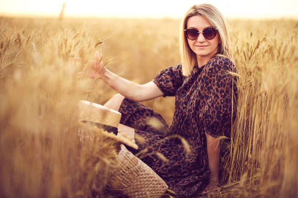 woman in animal print dress, straw hat in front of Sun in middle of wheat field with pleasure , enjoying summer holidays