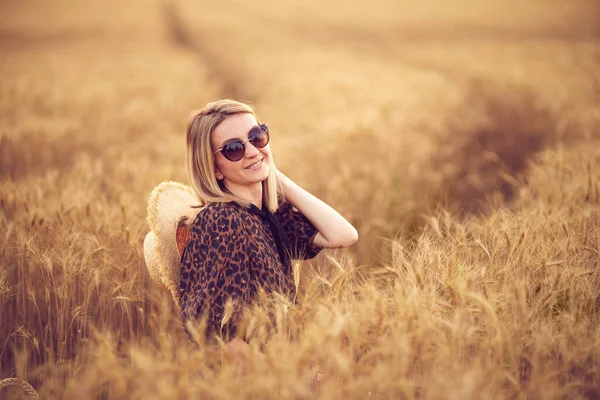 Mujer en vestido de animal print, sombrero de paja delante del sol en medio del campo de trigo con placer, disfrutando de las vacaciones de verano — Foto de Stock