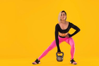 Şık, pembe ve siyah spor elbiseli kadın sarı arka planda kettlebell ile spor yapıyor..