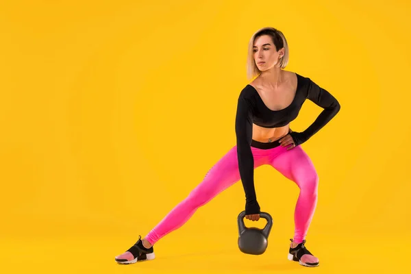 Mujer fitness en ropa deportiva rosa y negro de moda trabajar con kettlebell sobre fondo amarillo . — Foto de Stock