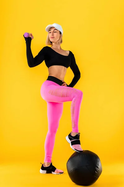 Αθλητική γυναίκα σε μοντέρνα αθλητικά ρούχα κάνει τις ασκήσεις με αλτήρες. Φωτογραφία της μυϊκής γυναίκας σε κίτρινο φόντο. — Φωτογραφία Αρχείου