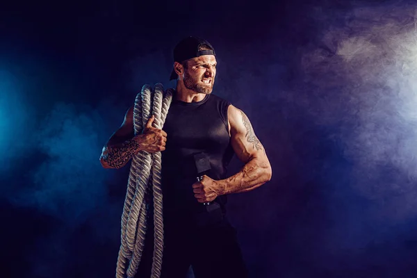 Brodaty atletyczny wyglądający bodybulder trzymający linę bojową i sztangę na ciemnym tle studio z dymem. Siła i motywacja — Zdjęcie stockowe