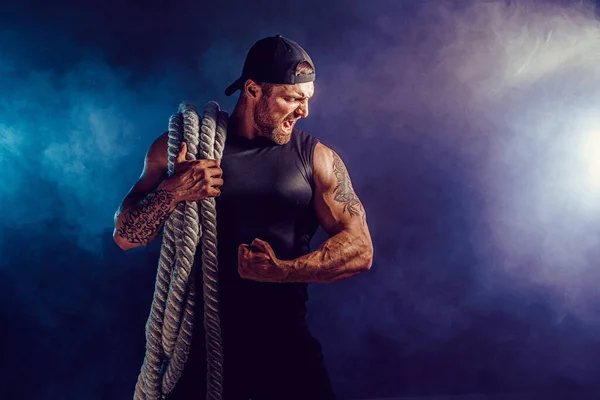 Bearded athletic looking bodybulder holding battle rope on dark studio background with smoke. Strength and motivation — Stock Photo, Image