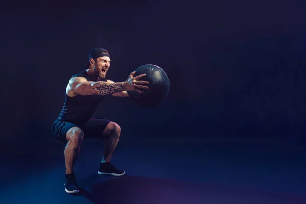 Aggressive bearded muscular sportsman is working out with a medicine ball isolated on dark studio background — Stock Photo, Image