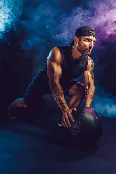 Agresywny, brutalny, brodaty, muskularny sportowiec ćwiczy z kulą lekarską odizolowaną na ciemnym tle studia z dymem — Zdjęcie stockowe