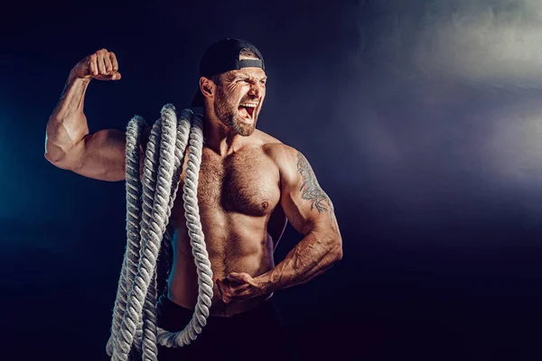 Aggressive bearded muscular sportsman holding battle rope on dark studio background with smoke. — Stock Photo, Image