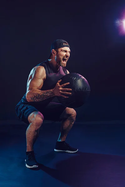 Aggressive bearded muscular sportsman is working out with a medicine ball isolated on dark studio background — Stock Photo, Image