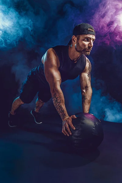 Aggressive bearded muscular sportsman is working out, push up with a medicine ball isolated on dark studio background with smoke