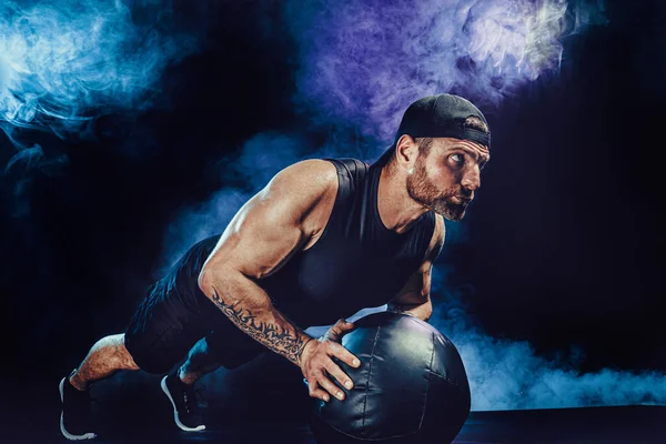 Агресивний бородатий м'язистий спортсмен працює, штовхаючи лікувальний м'яч ізольовано на темному студійному фоні з димом — стокове фото