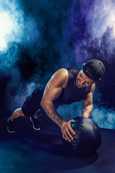 Aggressive bearded muscular sportsman is working out, push up with a medicine ball isolated on dark studio background with smoke — Stock Photo, Image