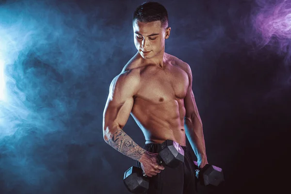 Atletický muž trénuje svaly s činkami ve studiu na tmavém pozadí s kouřem. Silný kulturista se šesti balení, perfektní svaly, ramena, biceps, triceps a hrudník — Stock fotografie