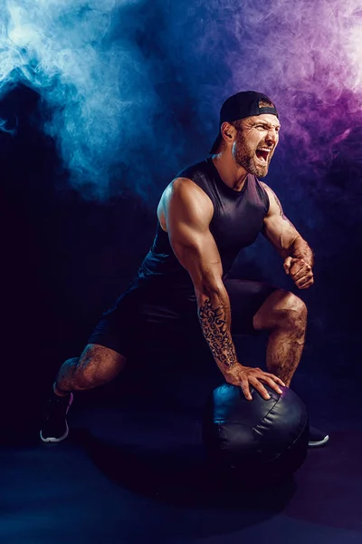 Aggressive bearded muscular sportsman is working out, push up with a medicine ball isolated on dark studio background with smoke — Stock Photo, Image