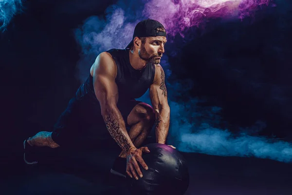 Agresivo barbudo musculoso deportista está trabajando, empujar hacia arriba con una pelota de medicina aislado en oscuro estudio fondo con humo — Foto de Stock