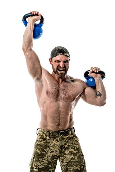 Muskulös idrottare bodybuilder man i camouflage byxor med en naken bål träning med kettlebell på en vit bakgrund. — Stockfoto