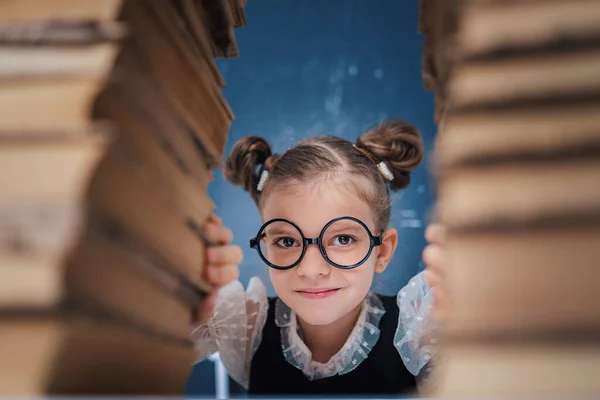 Щаслива розумна дівчина в округлих окулярах сидить між двома кулями книг і дивиться на камеру посміхаючись — стокове фото