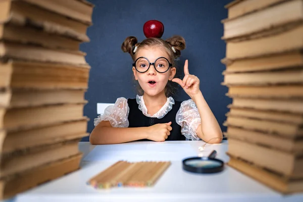 Menina inteligente feliz em óculos arredondados cuidadosamente sentado entre duas pilhas de livros com maçã vermelha na cabeça, apontando o dedo e olhar para a câmera sorrindo — Fotografia de Stock
