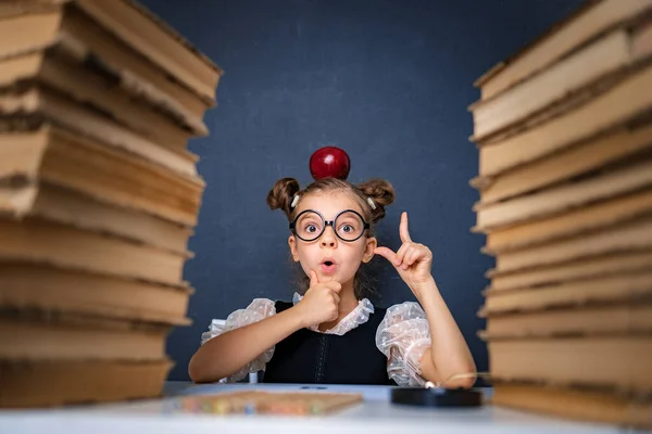 Menina inteligente feliz em óculos arredondados cuidadosamente sentado entre duas pilhas de livros com maçã vermelha na cabeça, apontando o dedo e olhar para a câmera sorrindo — Fotografia de Stock