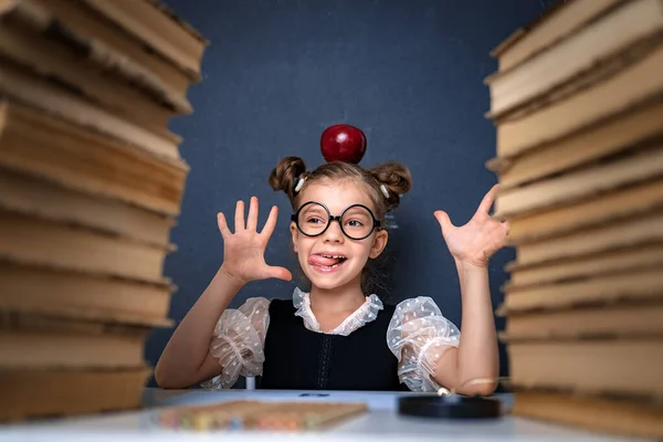 Glückliches intelligentes Mädchen in runden Gläsern mit rotem Apfel auf dem Kopf, das zwischen zwei Bücherstapeln sitzt, Spaß hat und in die Kamera lächelt — Stockfoto