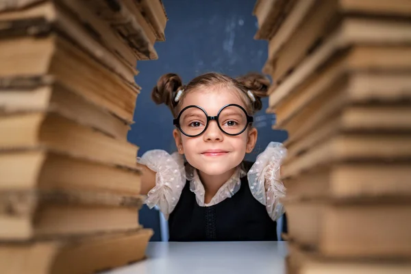 Ευτυχισμένο έξυπνο κορίτσι με στρογγυλά γυαλιά κάθεται ανάμεσα σε δύο σωρούς από βιβλία και να δούμε κάμερα χαμογελώντας — Φωτογραφία Αρχείου