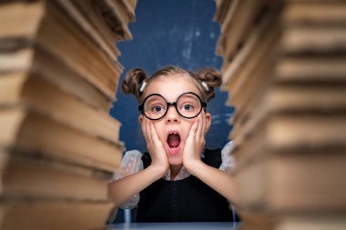 Yuvarlak gözlüklü mutlu akıllı kız iki kitap yığınının arasında oturuyor ve kameraya bak şaşırmış.