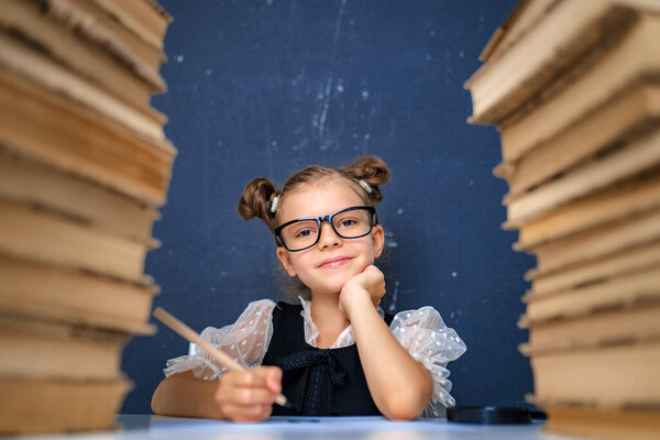 Happy smart girl in rounded glasses, holding a pencil in hand ready to write sitting between two piles of books and look at camera smiling