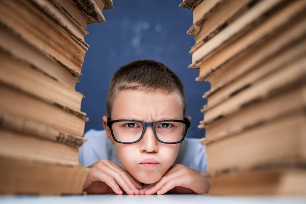 Az iskolás fiú szemüvegben ül két halom könyv között, és nézi a kamerát, ahogy kidagad az arca. — Stock Fotó