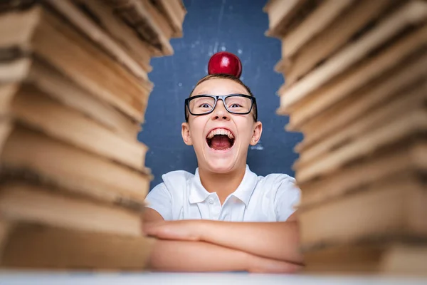 Glücklicher smarter Junge in Brille sitzt zwischen zwei Bücherstapeln mit rotem Apfel auf dem Kopf und blickt mit offenem Mund in die Kamera — Stockfoto