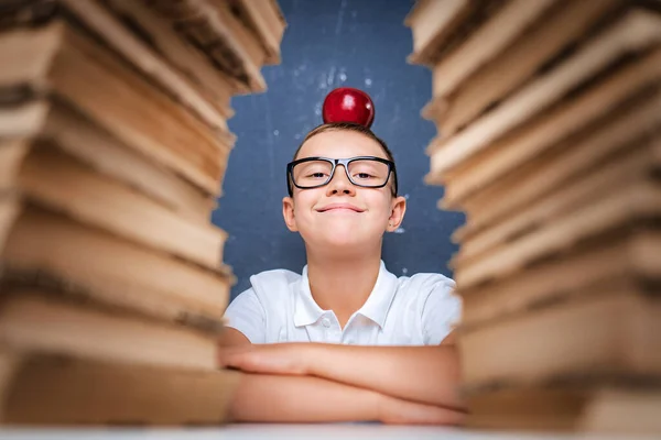 Boldog okos fiú szemüvegben ül két halom könyv között vörös almával a fején, és nézi a kamerát mosolyogva. — Stock Fotó