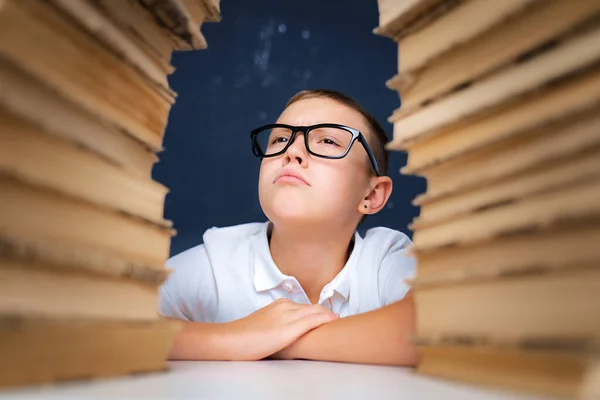 Kluger Junge mit Brille sitzt zwischen zwei Bücherstapeln und blickt aufgeblasene Wangen hoch — Stockfoto