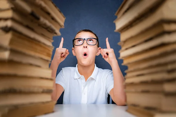 Kluger Junge mit Brille sitzt zwischen zwei Bücherstapeln und schaut auf, zeigt mit dem Finger — Stockfoto