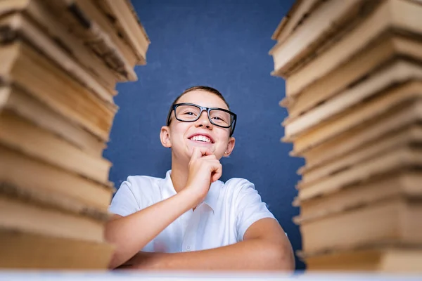 Okos fiú szemüvegben ül két halom könyv között, és figyelmesen felnéz. — Stock Fotó