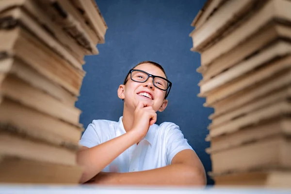 Boldog okos fiú szemüvegben ül két halom könyv között és mosolyogva néz fel. — Stock Fotó