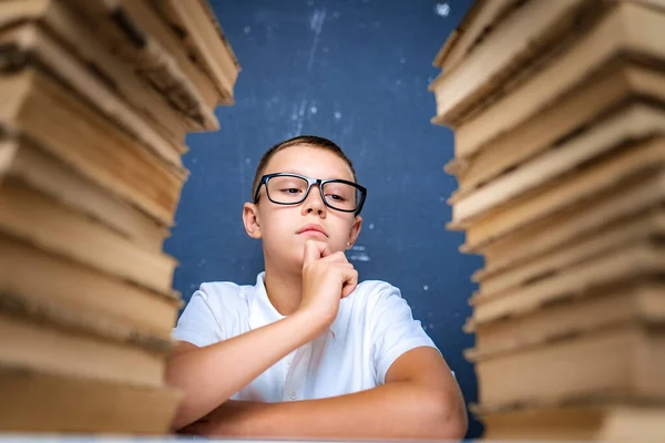 Kluger Junge mit Brille sitzt zwischen zwei Bücherstapeln und blickt nachdenklich nach unten — Stockfoto