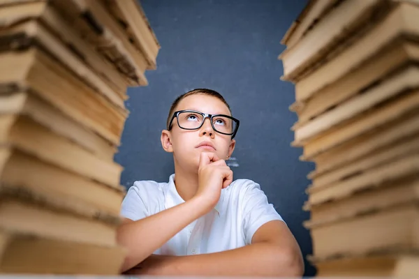 Okos fiú szemüvegben ül két halom könyv között, és figyelmesen felnéz. — Stock Fotó