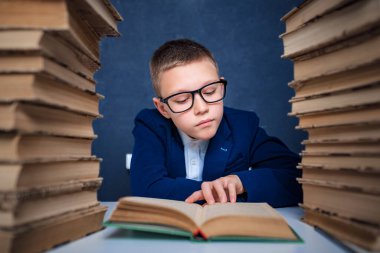 Gözlüklü zeki çocuk iki kitap yığınının arasında oturuyor ve dikkatlice kitap okuyor.