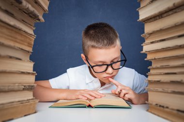 Gözlüklü zeki çocuk iki kitap yığınının arasında oturuyor ve dikkatlice kitap okuyor.