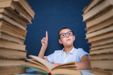 Gözlüklü zeki çocuk iki kitap yığınının arasında oturuyor ve yukarı bakıyor, işaret ediyor.