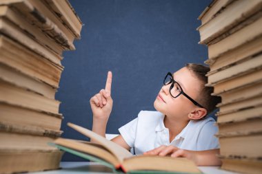 Gözlüklü zeki çocuk iki kitap yığınının arasında oturuyor ve yukarı bakıyor, işaret ediyor.