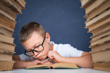 Gözlüklü zeki çocuk iki kitap yığınının arasında oturuyor ve dikkatlice kitap okuyor.