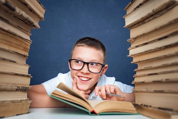 School boy in glasses sitting between two piles of books and look away from camera smiling — Stock Photo, Image