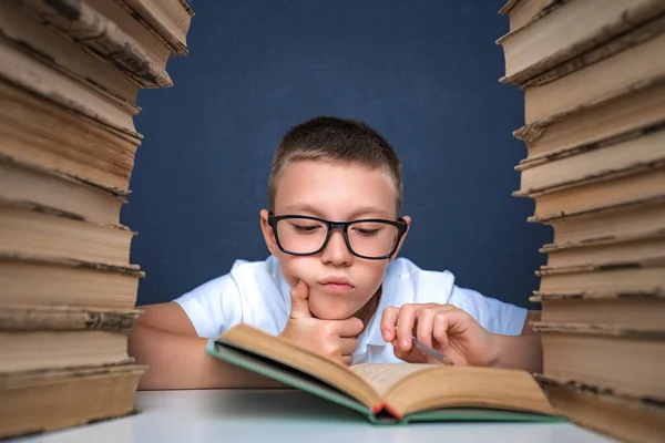 Kluger Junge mit Brille sitzt zwischen zwei Bücherstapeln und liest nachdenklich Buch — Stockfoto