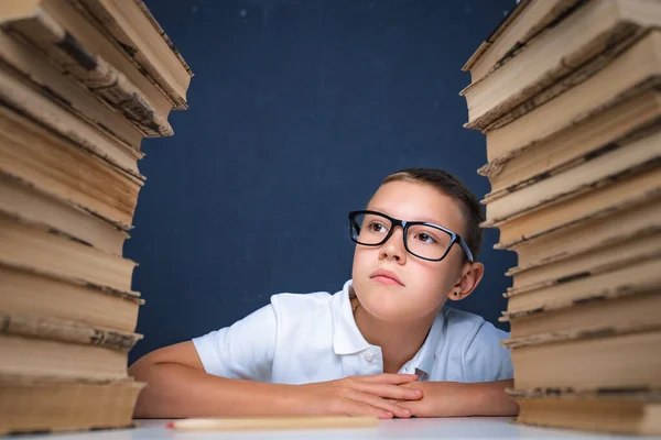 Έξυπνο αγόρι με γυαλιά που κάθεται ανάμεσα σε δύο σωρούς βιβλίων και κοιτάζει αλλού με προσοχή. — Φωτογραφία Αρχείου
