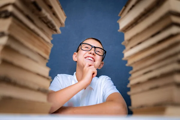 Glücklicher smarter Junge mit Brille sitzt zwischen zwei Bücherstapeln und blickt lächelnd nach oben — Stockfoto