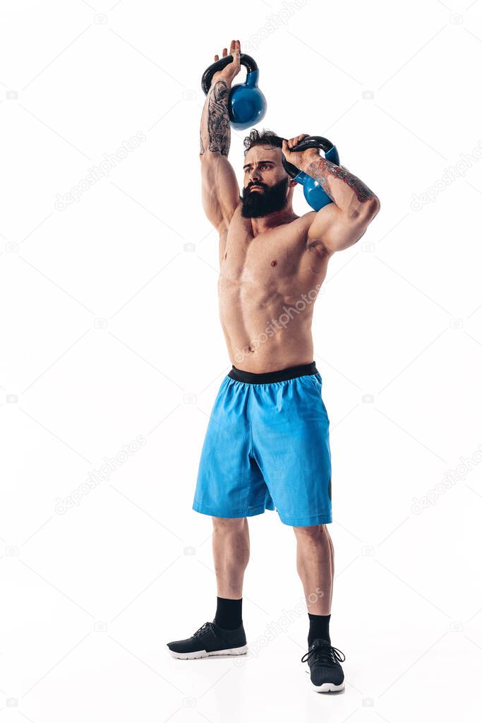 Muscular shirtless tattooed bearded male athlete bodybuilder workout with kettlebell on a white background.