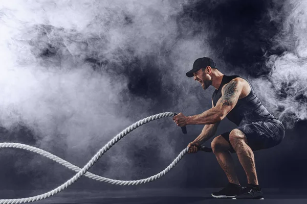 Γενειοφόρος αθλητική εμφάνιση bodybulder ασκηθείτε με σχοινί μάχης σε σκούρο φόντο στούντιο με καπνό. Δύναμη και κίνητρο — Φωτογραφία Αρχείου