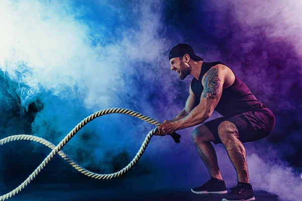 Γενειοφόρος αθλητική εμφάνιση bodybulder ασκηθείτε με σχοινί μάχης σε σκούρο φόντο στούντιο με καπνό. Δύναμη και κίνητρο — Φωτογραφία Αρχείου