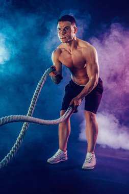 Atletik görünüşlü vücut bulucu, savaş halatıyla karanlık stüdyo arka planında dumanla çalışıyor. Kuvvet ve motivasyon