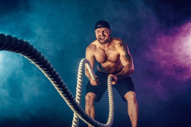 Sakallı, atletik görünümlü vücut bulucu. Karanlık stüdyo arka planında savaş halatıyla dumanla çalışıyor. Kuvvet ve motivasyon